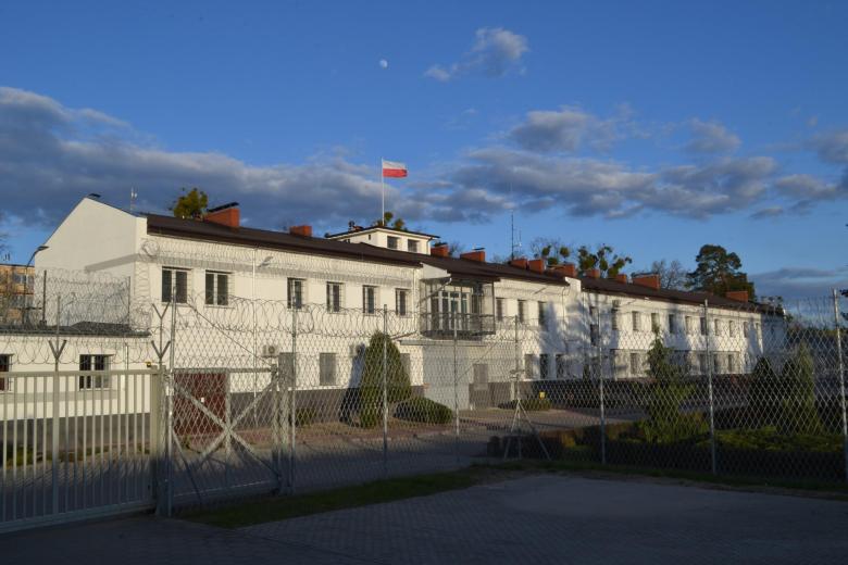 Dwupiętrowy, długi budynek za drutem kolczastym. Na szczycie budynku jest maszt z powiewającą biało-czerwoną flagą