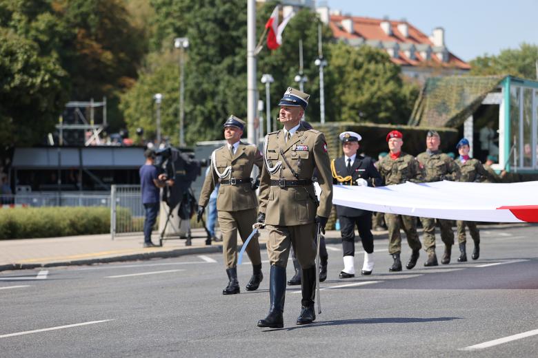 Oficer w mundurze wojskowym prowadzi defiladę, trzymając szablę. Za nim maszerują żołnierze w mundurach poszczególnych rodzajów sił zbrojnych i niosą wielką, biało czerwoną flagę.