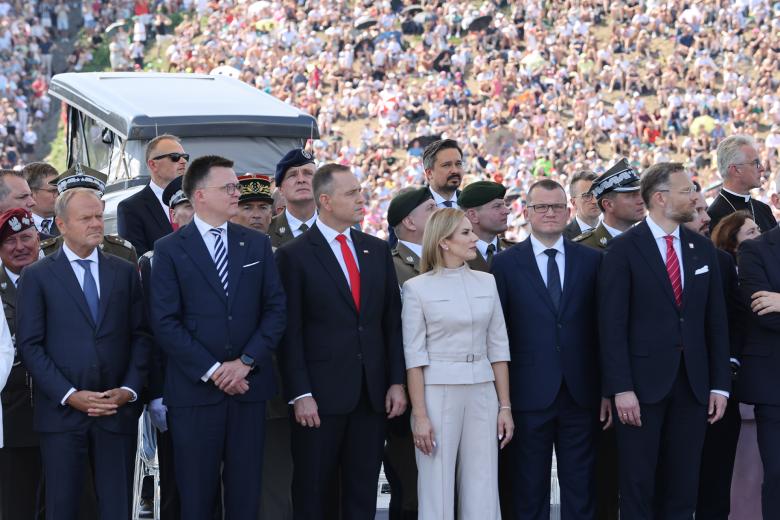 Kilkanaście osób, niektóre w garniturach, a niektóre w mundurach, stoi w trzech rzędach z głowami zwróconymi w stronę defilady. W tle widać tłum ludzi obserwujących defiladę.