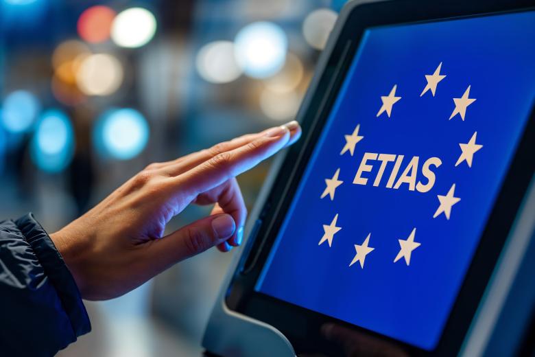 Osoba dotykająca palcem panelu dotykowego na którym na niebieskim tle jest widoczny żółty napis "ETIAS" otoczony 12 gwiazdkami.