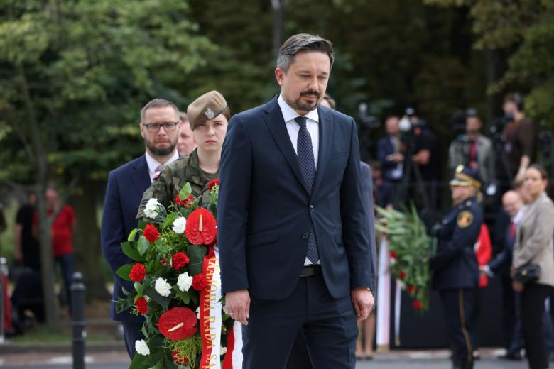 RPO Marcin Wiącek stoi wyprostowany, za nim stoi kobieta w mundurze, która trzyma wieniec