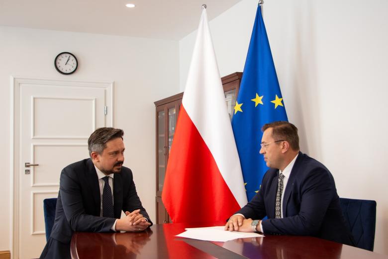 dwie osoby siedzą za stołem i rozmawiają na tle flag Polski i UE 