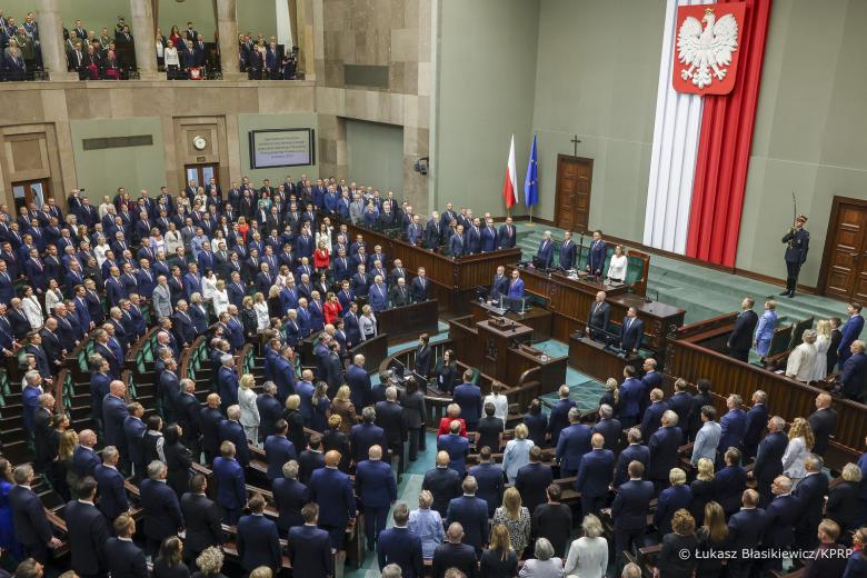 Pełna sala plenarna Sejmu, w której wszyscy zgromadzeni stoją. 