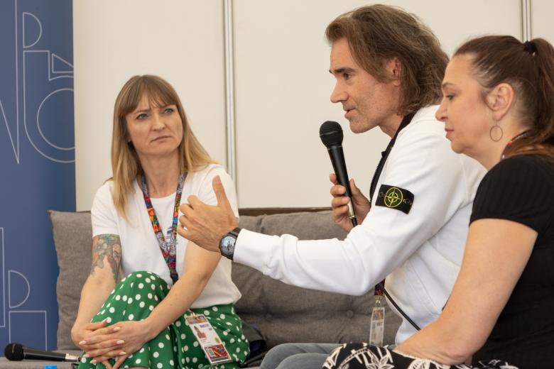 Trzy osoby siedzą obok siebie. Mężczyzna wypowiada się, trzymając w ręku mikrofon. Obok siedzą dwie kobiety i słuchają wystąpienia