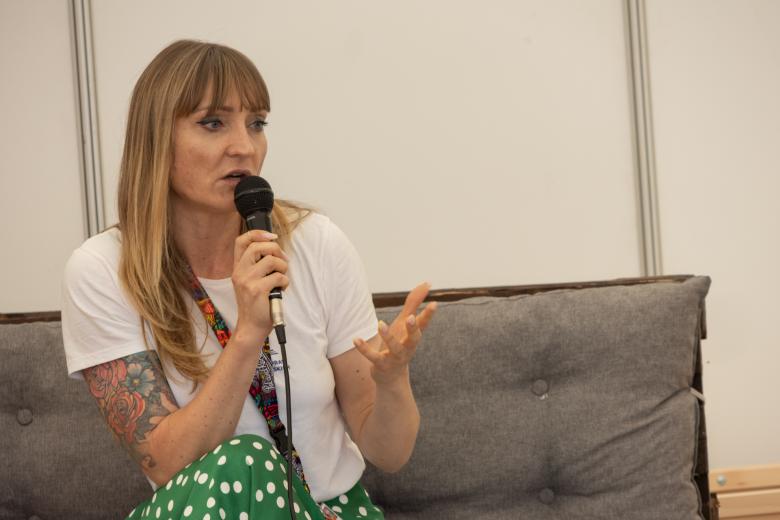 Kobieta ubrana w białą koszulkę i zieloną spódnicę w groszki siedzi, w ręku trzyma mikrofon i wypowiada się