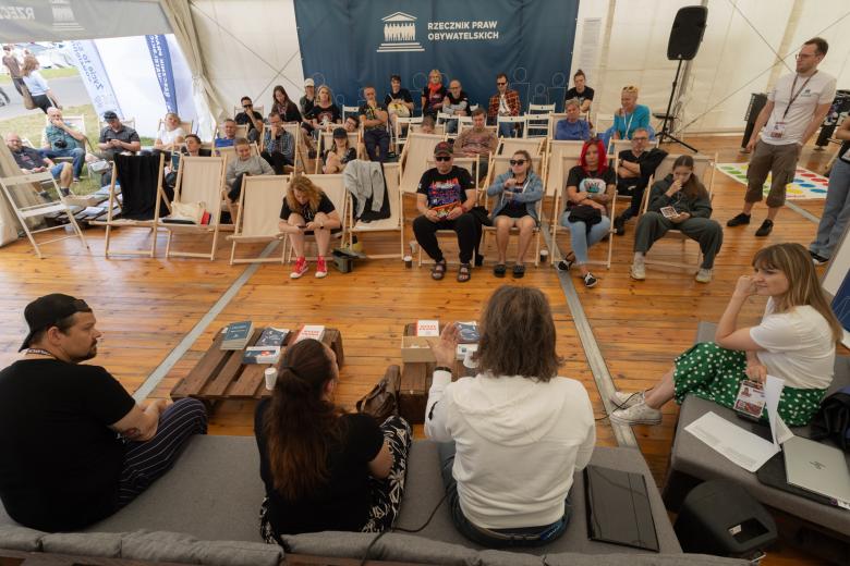 Grupa osób siedzi na leżakach i słucha czterech prelegentów, którzy siedzą przed nimi. W tle - na ścianie namiotu - widać napis "Rzecznik Praw Obywatelskich"