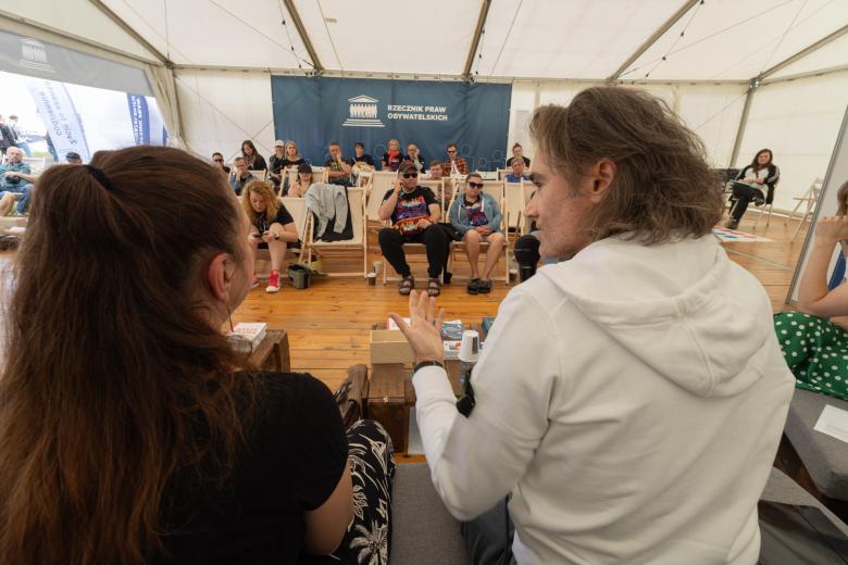 Dwie osoby siedzą obok siebie, jedna z nich trzyma w ręku mikrofon. Przed nimi siedzi kilkanaście osób i słucha 