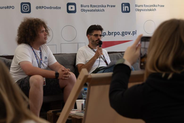 Dwie osoby siedzą obok siebie, jedna trzyma w ręku mikrofon i wypowiada się. Przed nimi są dwie osoby, jedna z nich robi zdjęcie telefonem