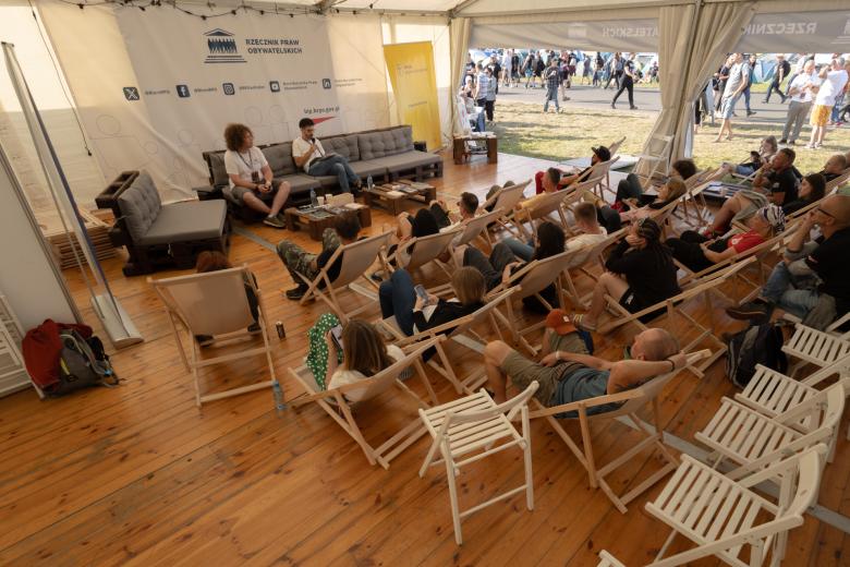 Kilkadziesiąt osób siedzi w namiocie na leżakach i krzesłach i słucha wystąpienia prelegentów