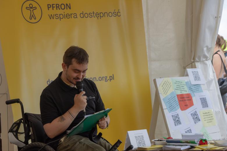 Mężczyzna siedzi na wózku, w ręku trzyma teczkę i mikrofon i wypowiada się. Za nim widać baner z napisem "PFRON wspiera dostępność" 