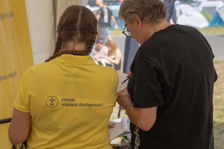 Dwie osoby stoją obok siebie tyłem, jedna ma na sobie żółtą koszulkę z napisem "PFRON wspiera dostępność", a druga czarny t-shirt 