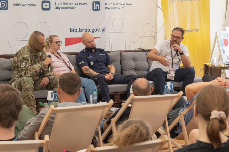 Czterech prelegentów siedzi obok siebie, dwóch z nich trzyma w ręku mikrofon i wypowiada się. Przed nimi na leżakach siedzą słuchacze