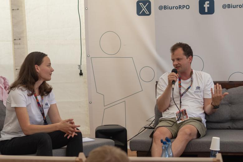 ZRPO Adam Krzywoń siedzi, trzyma w ręku mikrofon i wypowiada się. Obok siedzi kobieta i słucha jego wypowiedzi
