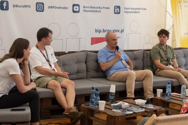 Cztery osoby siedzą, jedna z nich trzyma w ręku mikrofon i wypowiada się, pozostałe słuchają