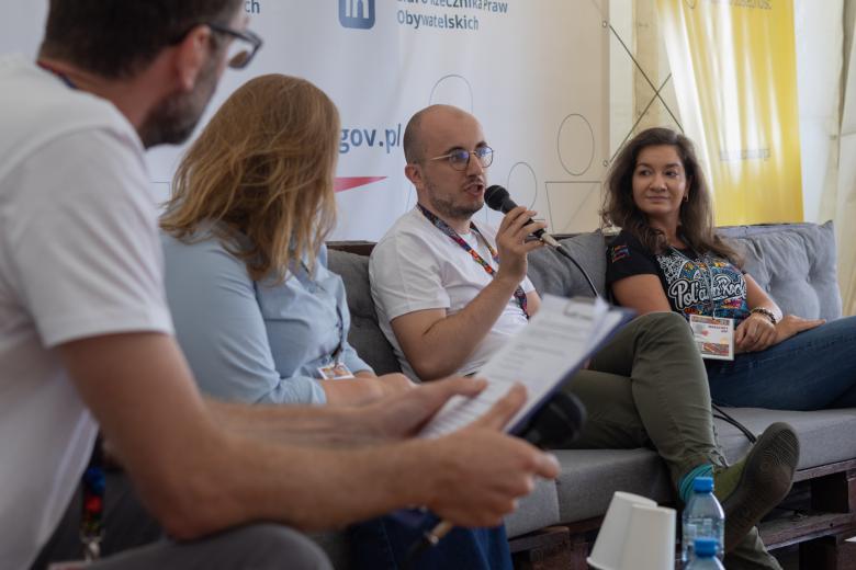 Cztery osoby siedzą obok siebie, jedna z nich trzyma w ręku mikrofon i wypowiada się, pozostałe słuchają