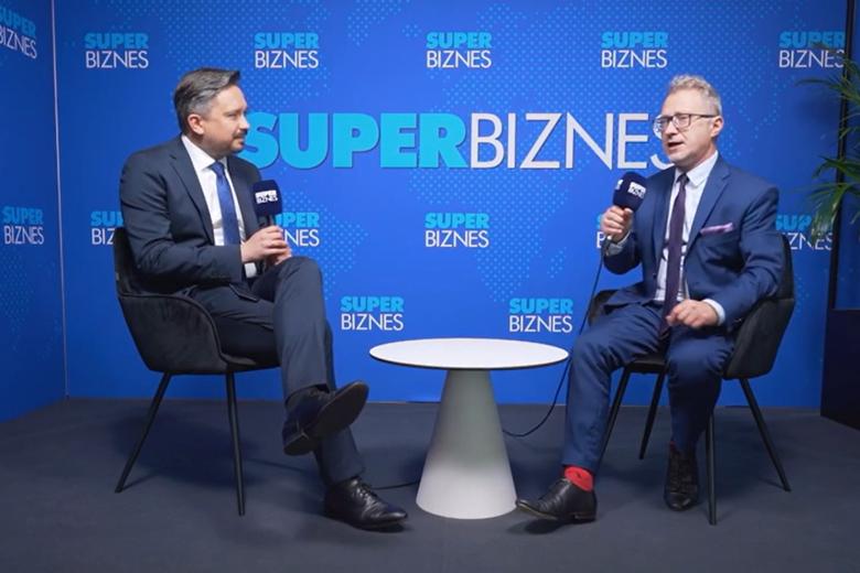 Dwie osoby siedzą na krzesłach przy stoliku i rozmawiają trzymając w dłoniach mikrofony, w tle ścianka z napisem "SuperBiznes"