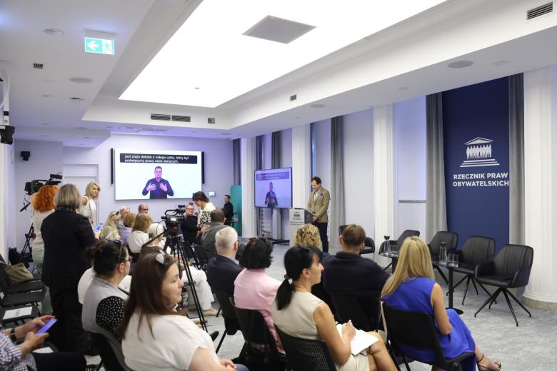 Kilkadziesiąt osób siedzi na sali konferencyjnej i słucha wystąpienia osoby mówiącej za mównicą na środku