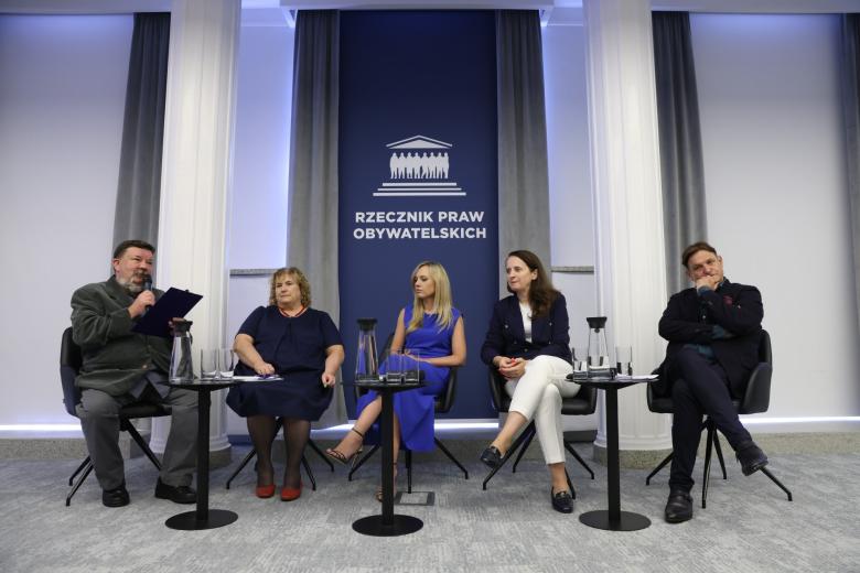 Pięć osób siedzi na krzesłach na sali konferencyjnej, jedna z nich mówi do trzymanego w dłoni mikrofonu