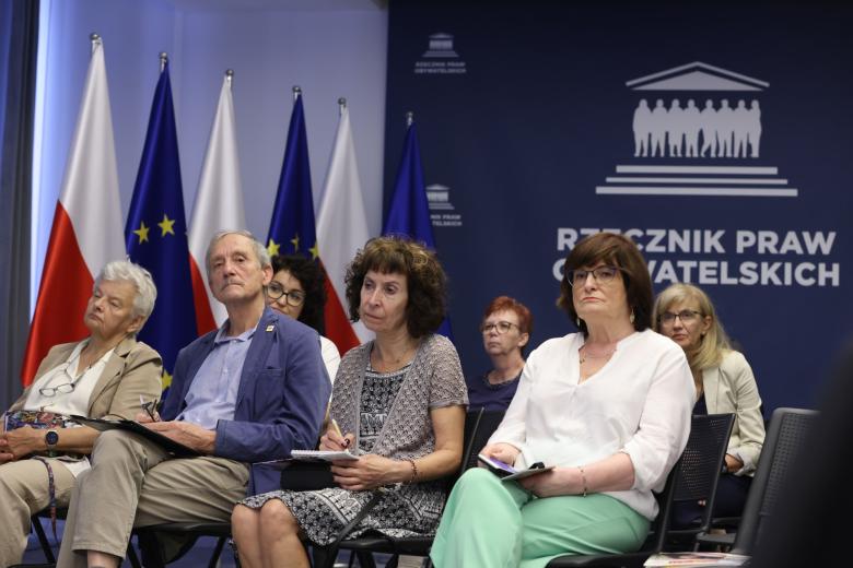 osoby siedzące na krzesłach z uwaga słuchają czyjegoś wystąpienia, w tle flagi Polski i UE oraz biały napis "Rzecznik Praw Obywatelskich" na granatowym tle