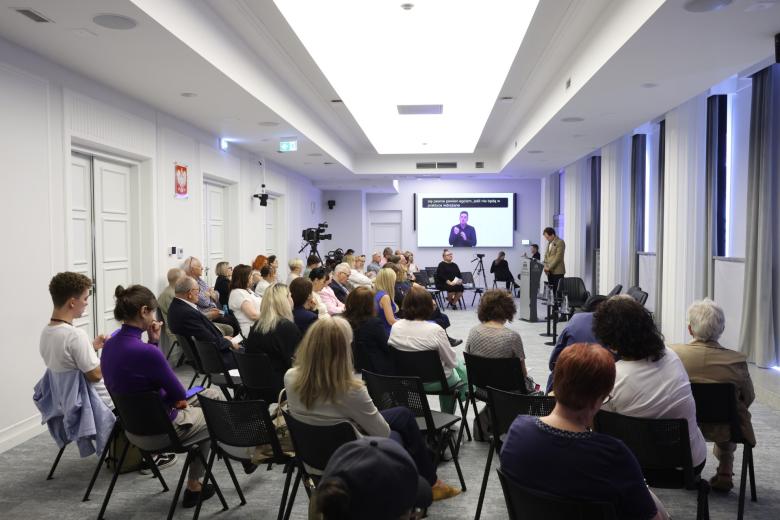 Kilkadziesiąt osób siedzi na sali konferencyjnej i słucha wystąpienia osoby mówiącej za mównicą na środku