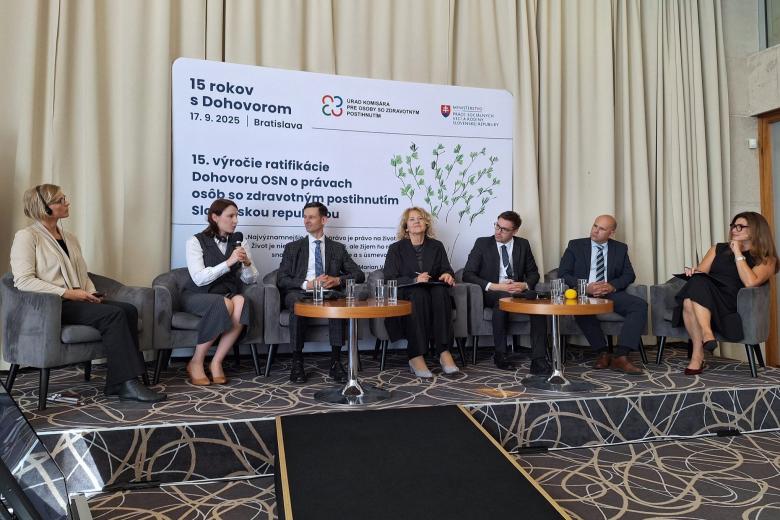 Siedem osób siedzi na krzesłach na podwyższeniu na sali konferencyjnej, jedna z nich mówi do trzymanego w dłoni mikrofonu