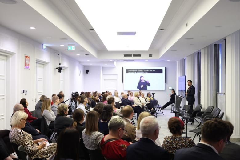 Kilkadziesiąt osób na sali konferencyjnej siedzi i słucha wystąpienia osoby mówiącej za mównicą