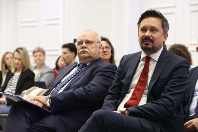 Dwie osoby siedzą na krzesłach na sali konferencyjnej i słuchają wystąpienia