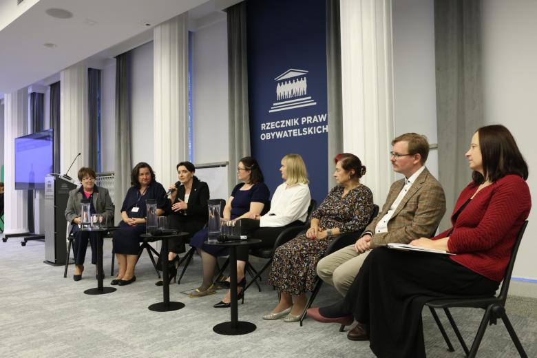 Osiem osób siedzi na krzesłach w sali konferencyjnej, jedna z nich trzyma w ręku mikrofon