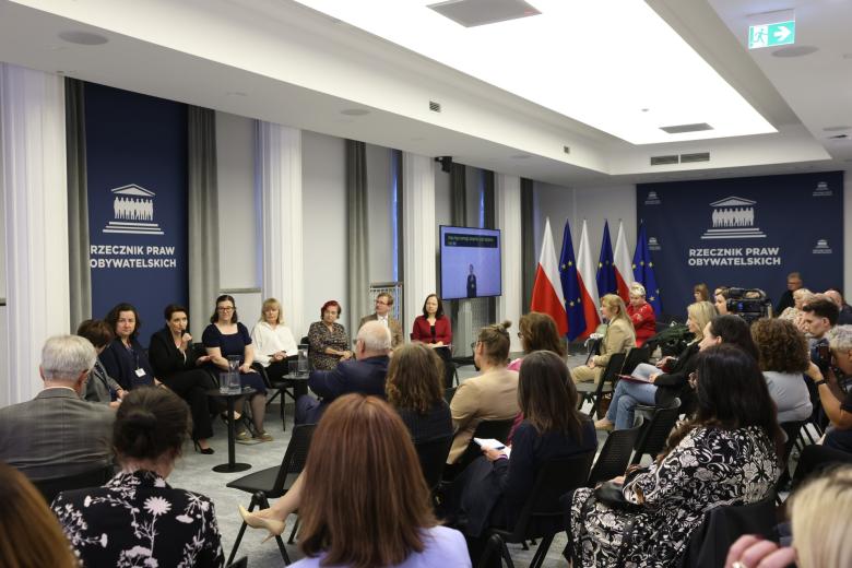 Kilkadziesiąt osób siedzi na sali konferencyjnej i słucha wystąpienia jednej z osób, które siedzą na krzesłach z przodu sali 