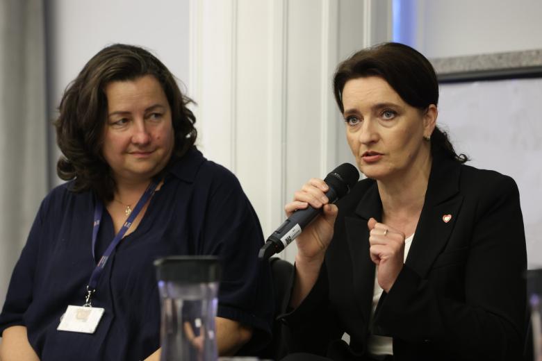 Dwie osoby siedzą na sali konferencyjnej, jedna z nich mówi do mikrofonu trzymanego w ręku