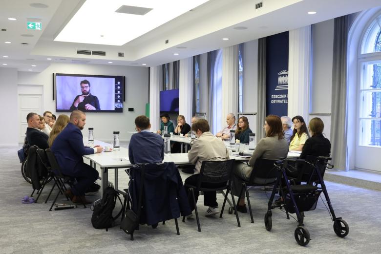 Kilkanaście osób siedzi przy stołach ustawionych w podkowę, na dużym telewizorze na ścianie widoczni uczestnicy posiedzenia łączący się zdalnie za pomocą programu do komunikowania się na odległość