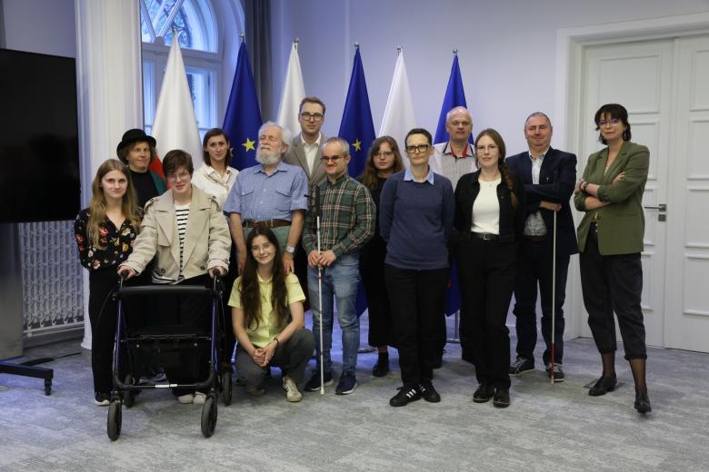 Kilkanaście osób pozuje na stojąco do zdjęcia na tle flag Polski i UE