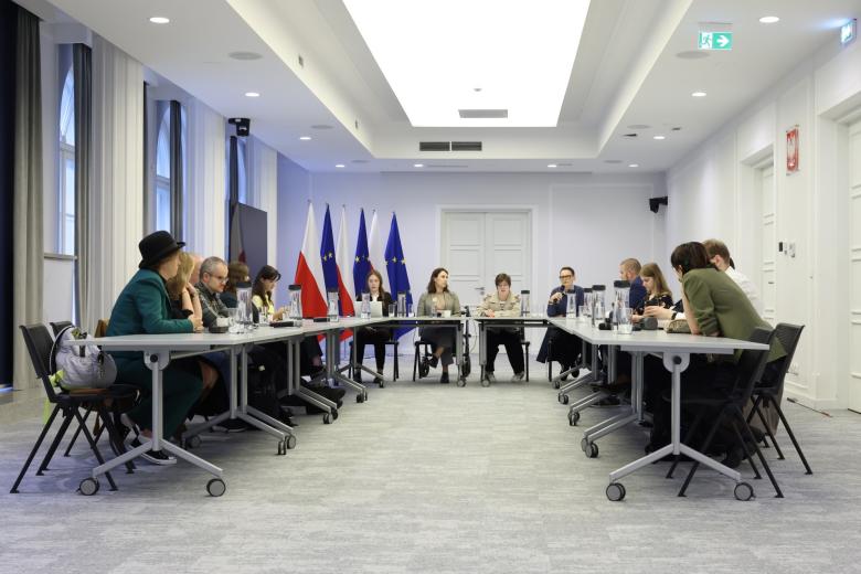 Kilkanaście osób siedzi przy stołach ustawionych w podkowę na sali konferencyjnej