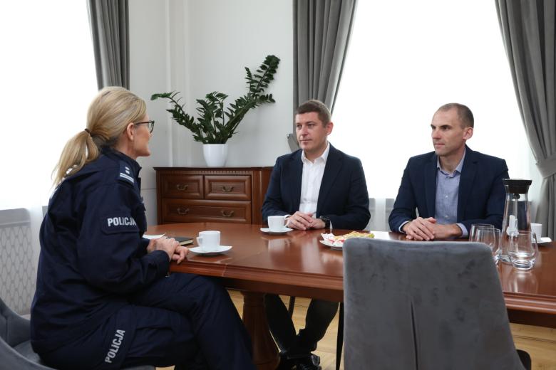 Trzy osoby, w tym jedna w mundurze policyjnym, siedzą przy stole i rozmawiają