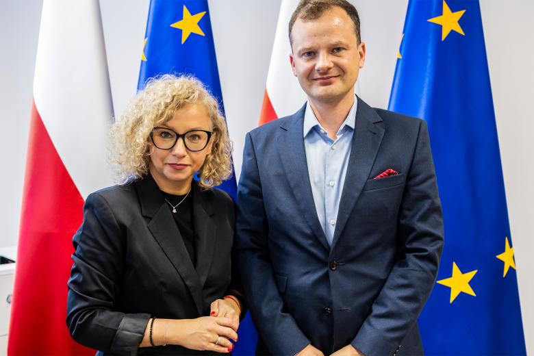 Dwie osoby pozują do wspólnego zdjęcia na tle flag Polski i UE