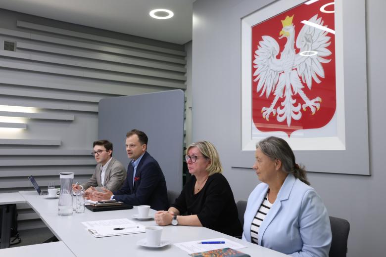 Cztery osoby siedzą za stołem w sali konferencyjnej i słuchają czyjegoś wystąpienia