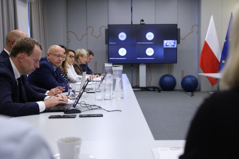 Kilka osób siedzi za podłużnym stołem w sali konferencyjnej, w tle wyświetlacz z uczestnikami spotkania łączącymi się na odległość