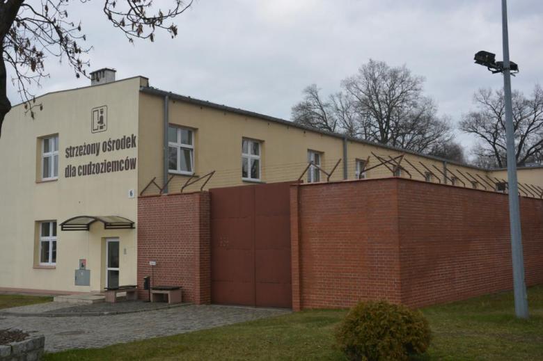 jednopiętrowy budynek z napisem "Strzeżony ośrodek dla cudzoziemców" z przylegającym wysokim murem z drutem kolczastym na górze