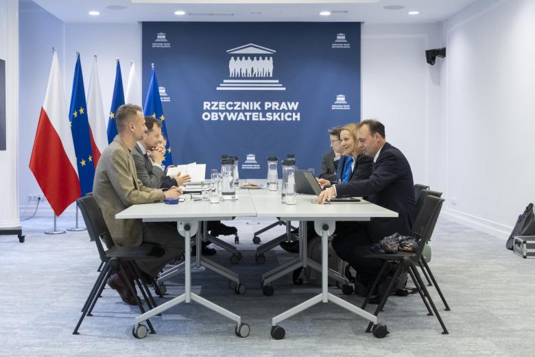 Kilka osób siedzi po dwóch stronach dużego prostokątnego stołu w sali konferencyjnej i rozmawia, w tle na dużej planszy napis "Rzecznik Praw Obywatelskich", a obok stojące flagi Polski i UE