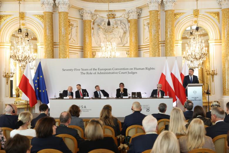Osoba przemawia za mównicą w bogato zdobionej zabytkowej sali. Obok mównicy stoi długi stół prezydialny, za którym siedzi 5 osób, w tle na białej planszy napis w języku angielskim "Seventy-Five Years of the European Convention on Human Rights: The Role of Administrative Court Judges"