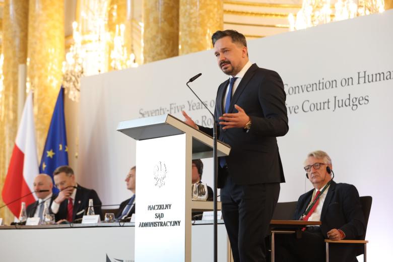 Osoba przemawia za mównicą w bogato zdobionej, zabytkowej sali