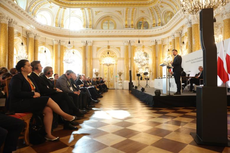 Kilkadziesiąt osób siedzących na krzesłach w bogato zdobionej, zabytkowej sali słucha wystąpienia osoby mówiącej za mównicą