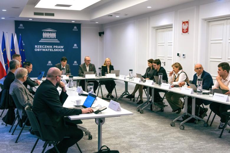 Kilkanaście osób siedzi przy stołach ustawionych w podkowę w sali konferencyjnej