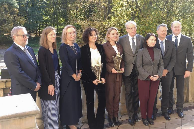 Grupa uśmiechniętych osób pozuje do zdjęcia. Dwie kobiety w środku grupy trzymają w dłoniach statuetki. 