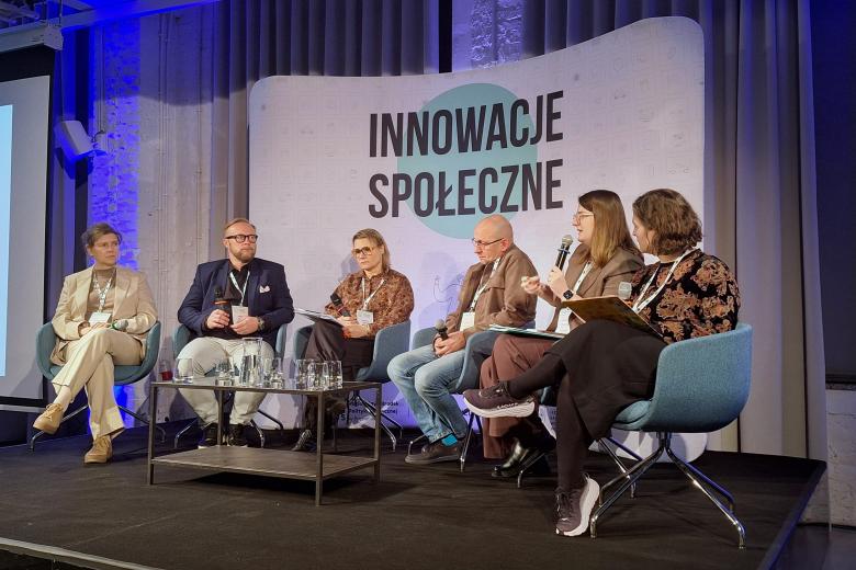 Sześć osób siedzi na scenie na krzesłach, jedna z nich mówi do mikrofonu, w tle napis "Innowacje Społeczne"