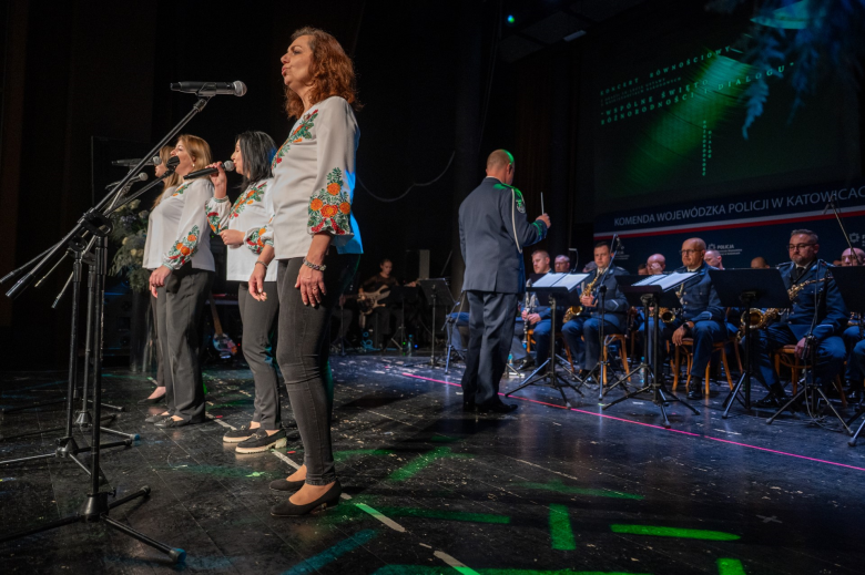 osoby śpiewają  na stojąco, w tle siedzą członkowie orkiestry 