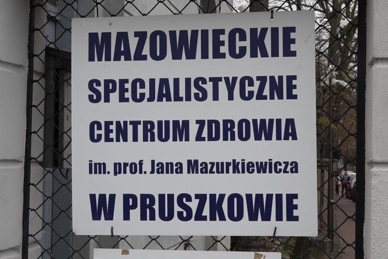 Tablica z napisem "Mazowieckie specjalistyczne centrum zdrowia im. prof. Jana Mazurkiewicza w Pruszkowie" zawieszona na ogrodzeniu
