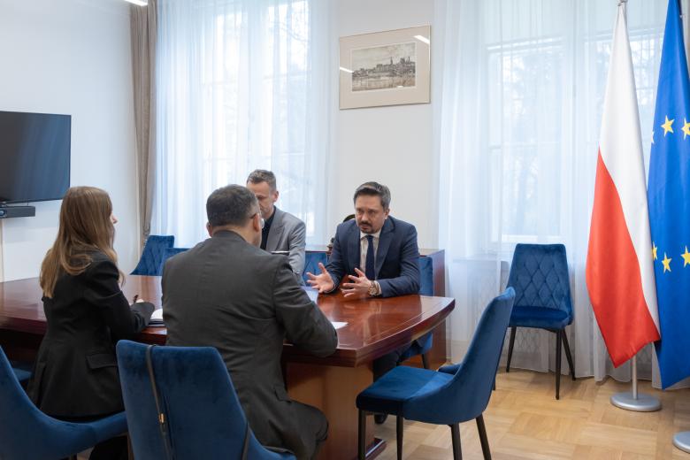 Cztery osoby siedzą przy stole i rozmawiają w salce konferencyjnej. Obok stołu stoją flagi Polski i Unii Europejskiej