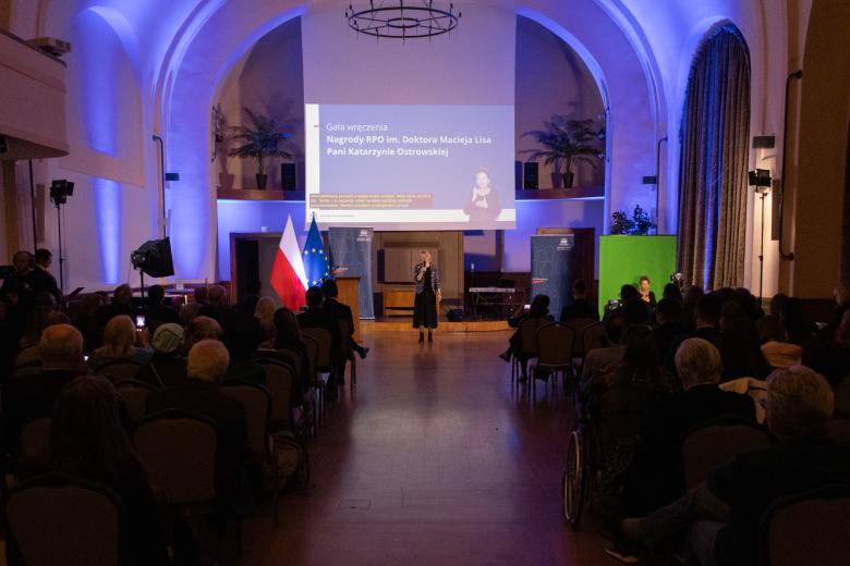 Katarzyna Ostrowska stoi przed publicznością na środku sali i wypowiada się do mikrofonu. 