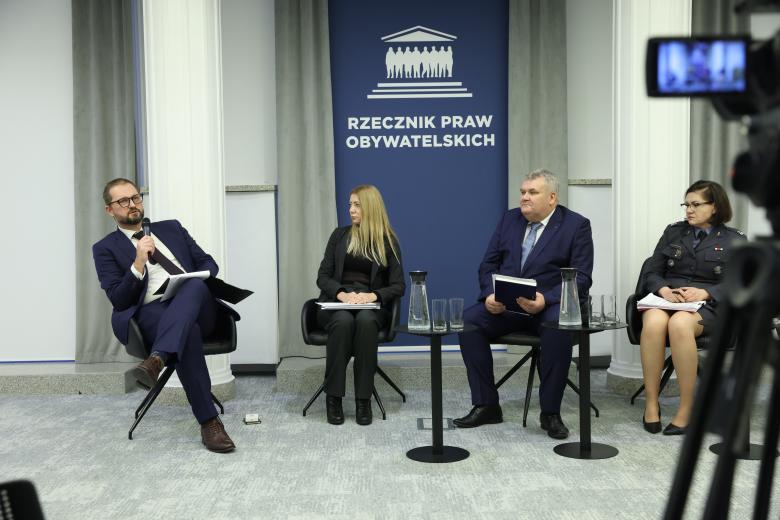 Cztery osoby rozmawiają siedząc obok siebie na krzesłach w sali konferencyjnej.